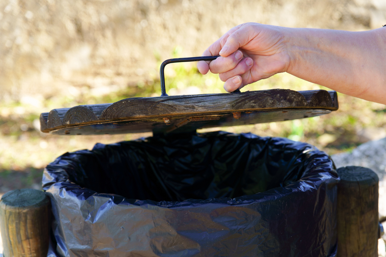 Human hand opening a rustic wooden garbage can with a black liner in an outdoor park setting for waste disposal